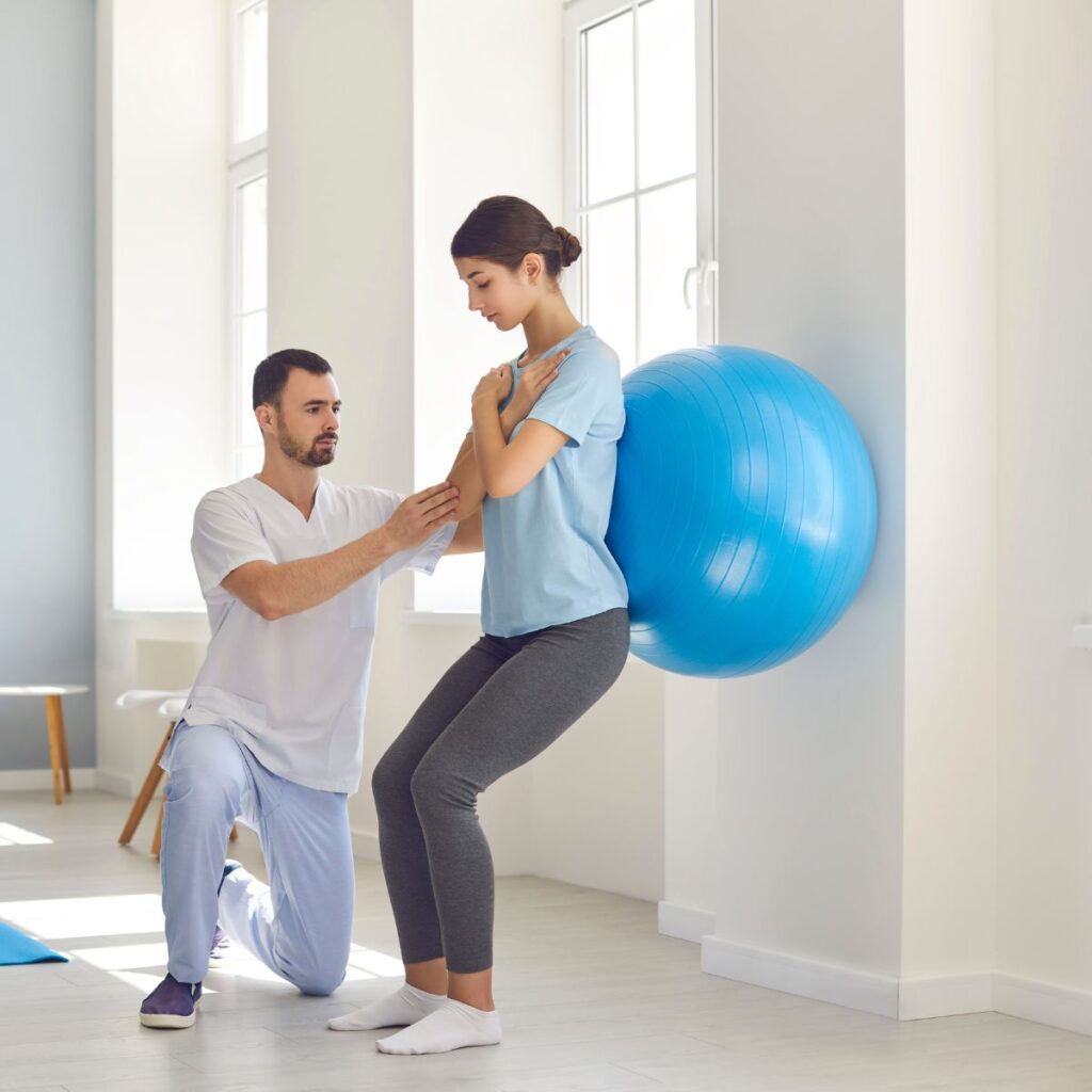 A physical therapist helps a woman do squats against a wall with an exercise ball