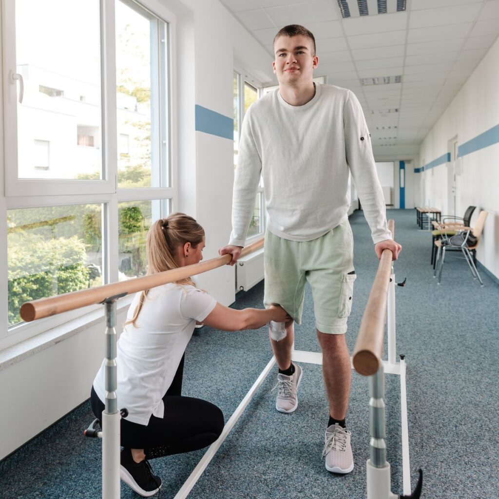 A therapist supports a young man practicing walking with parallel bars